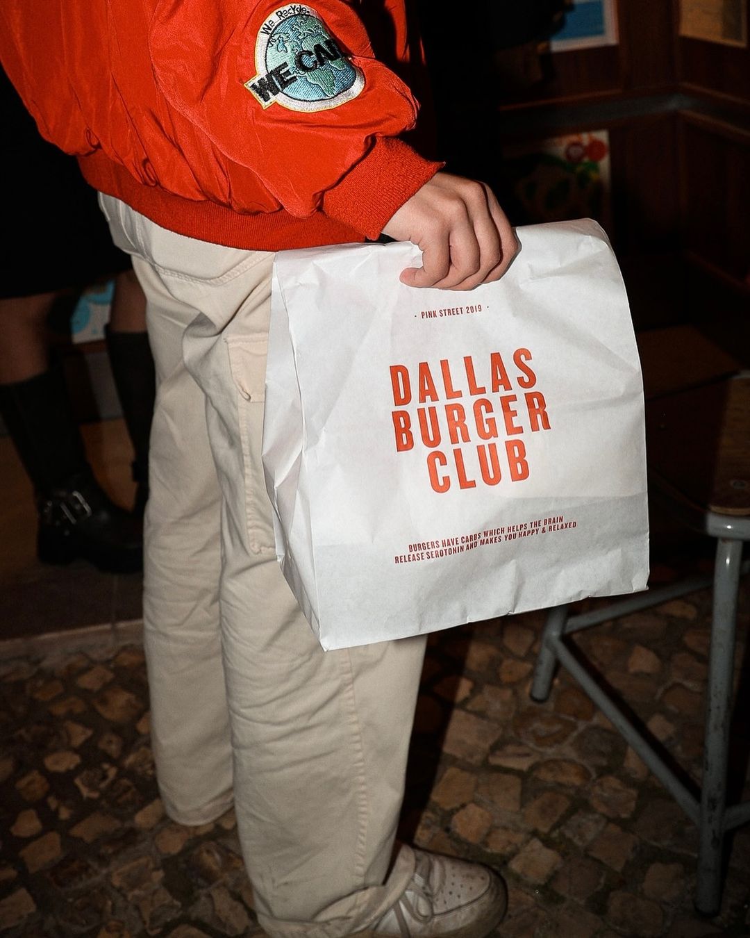 Person holding a Dallas Burger Club branded takeaway bag while wearing a bright orange jacket, standing on a cobblestone street at night